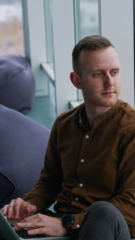 thoughtful man sit on windowsill in office. businessman with a laptop on laps looking at cityscape through panoramic window daydreaming about future. vertical video