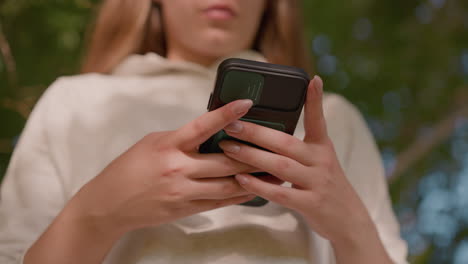 close-up of individual focus operating smartphone with polished nails, typing with precision while shadows fall softly on light-colored clothing, with a naturally blurred green background