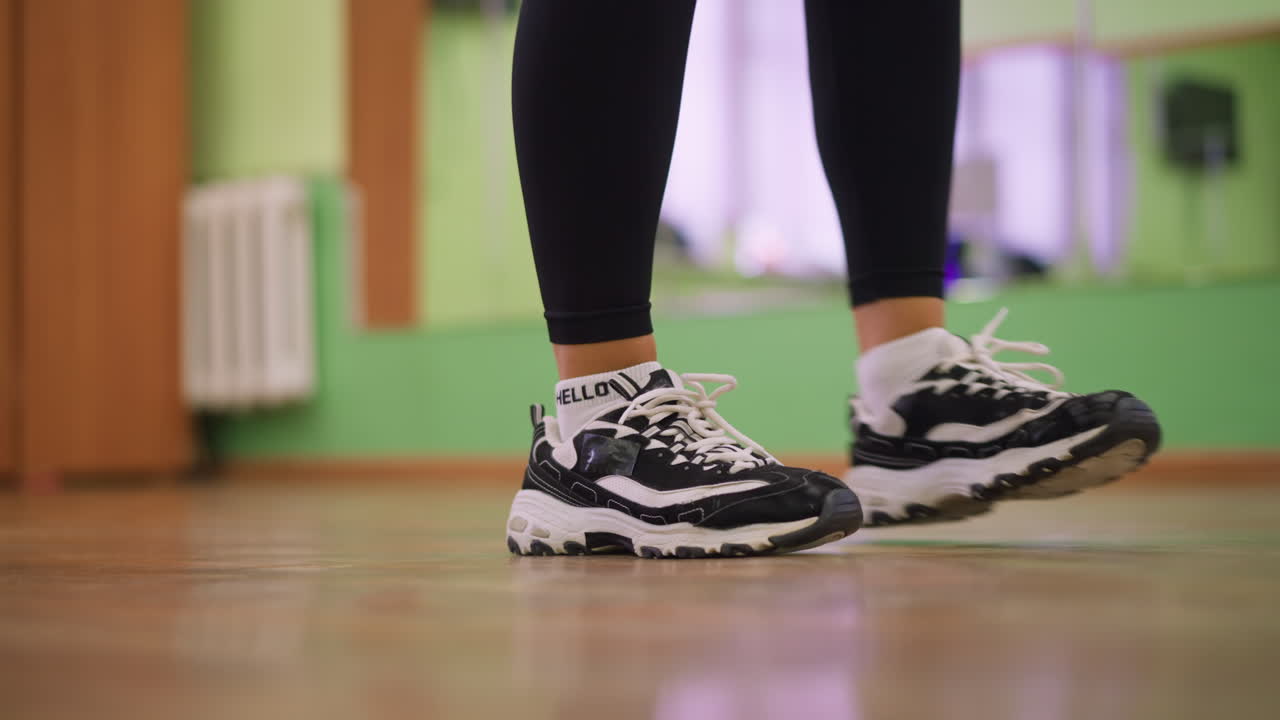 Leg view of girl in black leggings and canvas sneakers bouncing on polished wooden floor during indoor workout, showcasing rhythm, energy, and athletic movement in dynamic training session