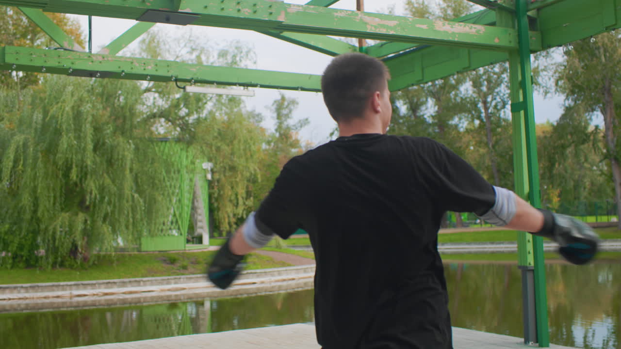 white male throwing wide punches outdoors under green metal pavilion by pond, explosive rotation and extended reach, black gloves, dynamic warmup, high energy motion, solo session focusing on speed