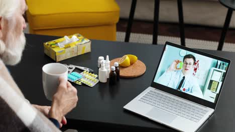 close up of sick man with cup of hot drink sitting in room and speaking on video on laptop with doctor