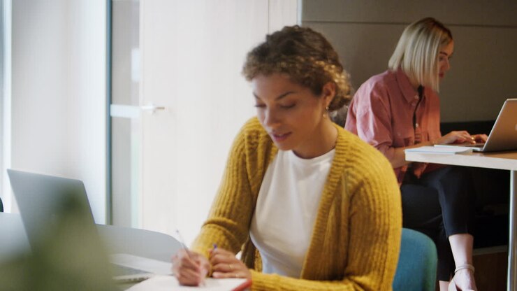 Smilende ung kvinde arbejder ved en bærbar computer på et kreativt kontor, mens hendes kollega arbejder i baggrunden.