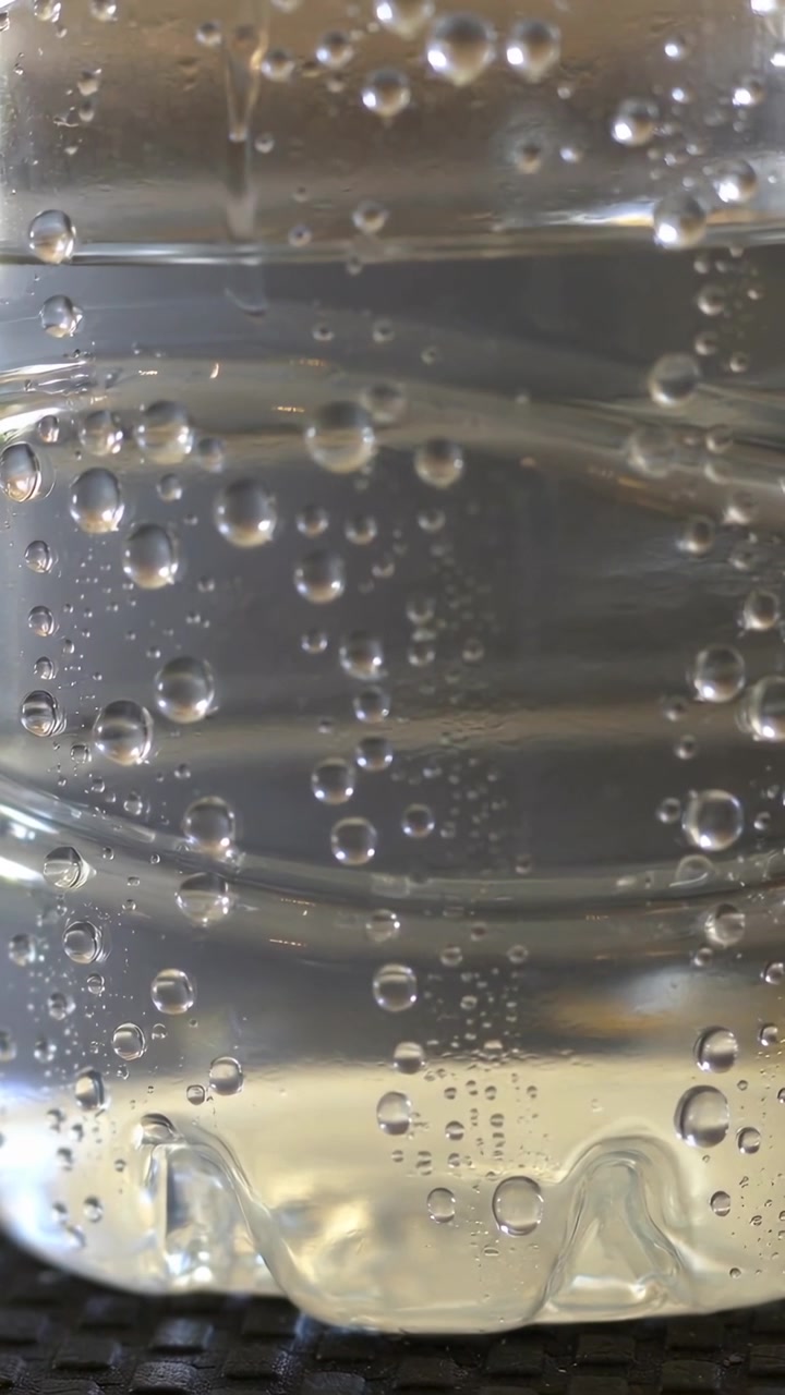 Video verticale: Mostra una bottiglia di plastica trasparente di acqua frizzante che forma e fa salire bolle su un tappetino scuro.