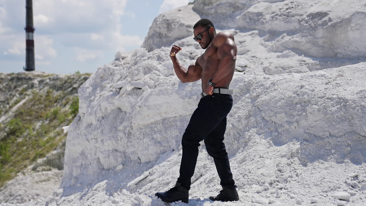 Muscular athlete on white rocky background. African american man in black glasses without shirt showing his biceps. Profile view.
