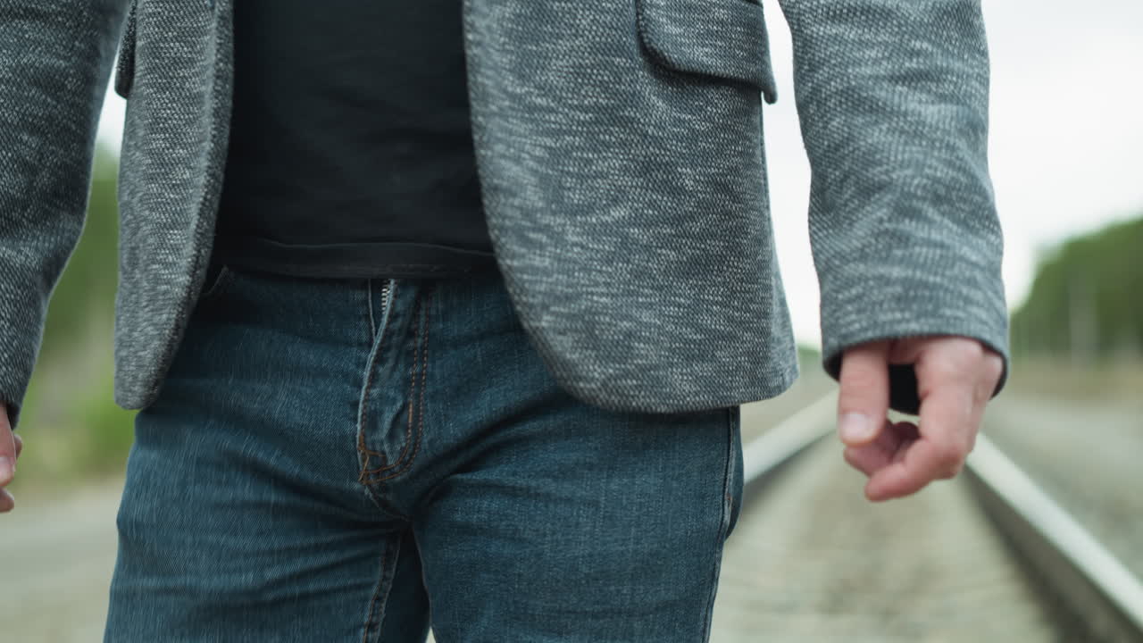 회색 슈트와 검은색 폴로 셔츠를 입은 남자가 철도 트랙을 고 주변이 흐릿한 모습으로 그의 옷에는 회색 블레이저와 파란 청바지가 포함되어 있습니다.