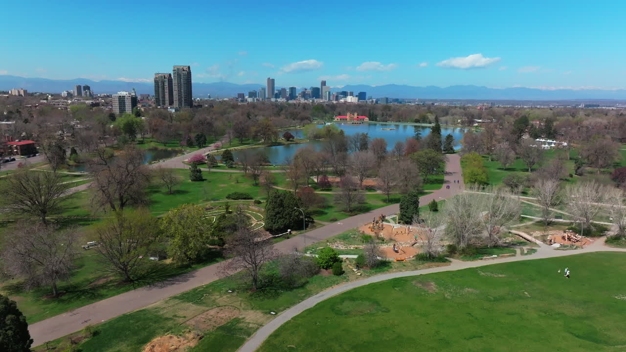 City Park Denver Colorado Lake Pavilion vibrant spring summer aerial drone sunny blue sky snow capped Rocky Mountains front range cityscape green lush grass trees blossom pan left motion