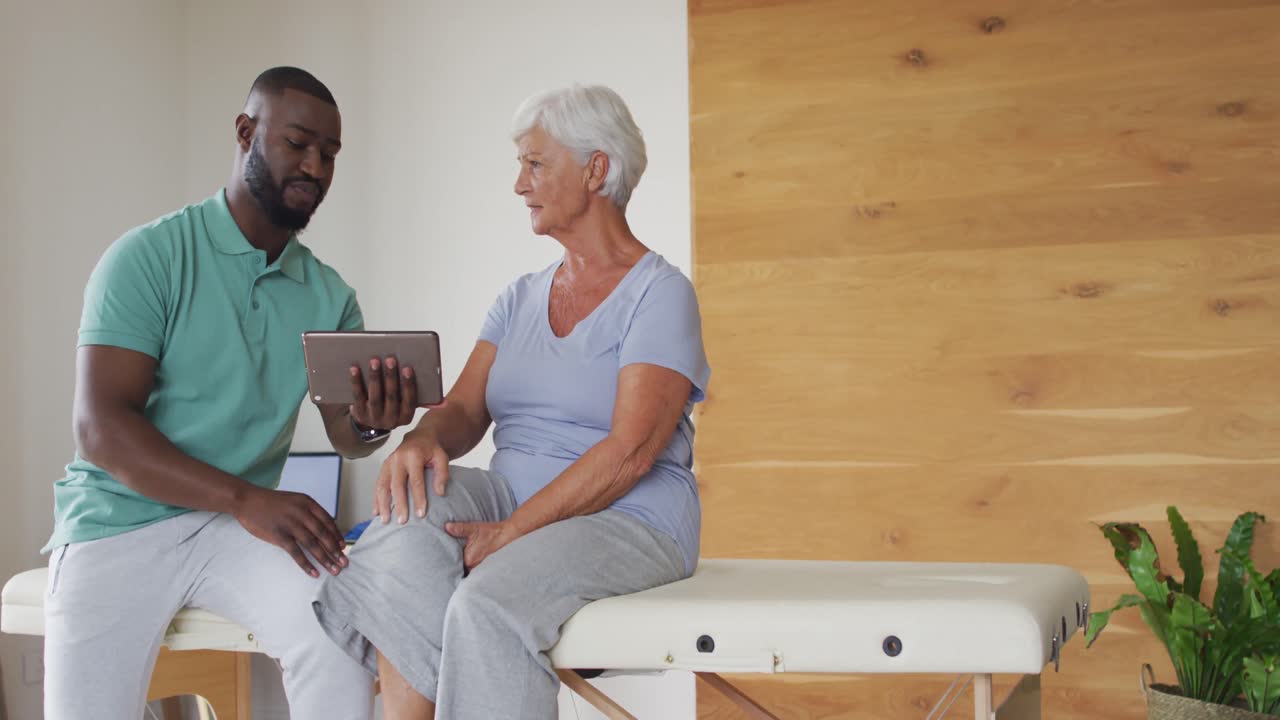 Video of african american male physiotherapist using tablet with caucasian senior woman
