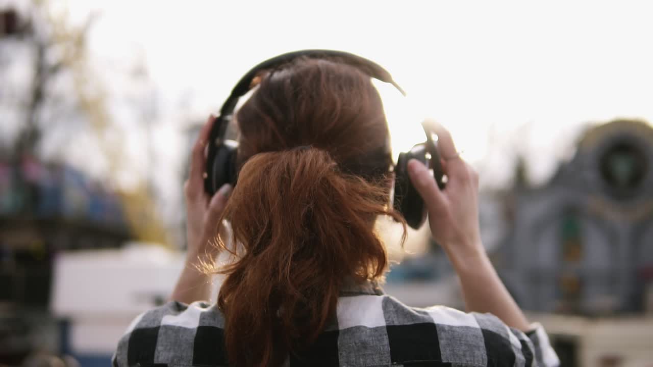 Backside footage of a girl with a brown haired tail standing a plaid shirt oover against the shining sun. Putting on the headphones