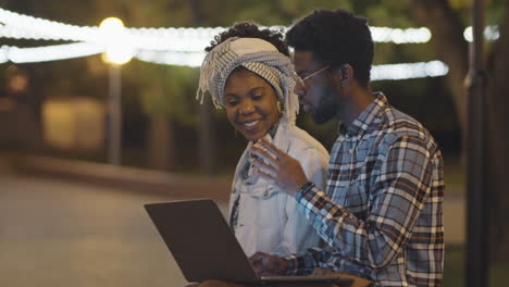 black man and woman using laptop and chatting in park in evening