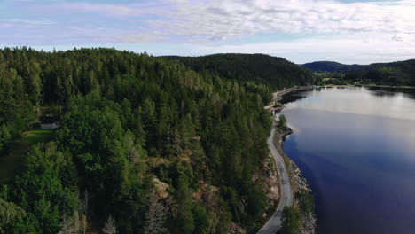 Aerial-view-of-narrow-road-between-calm-blue-lake-and-green-forest