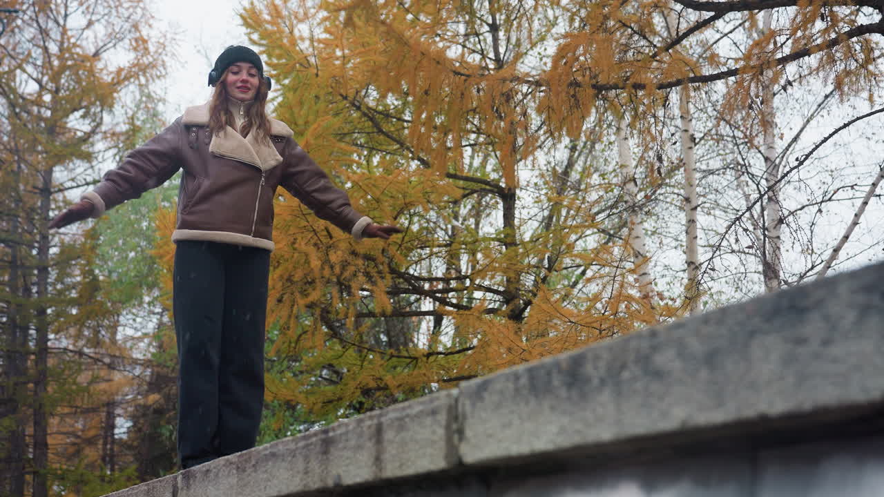 Smiling lady wearing black knit cap, brown shearling jacket, and black trousers gracefully balances with arms outstretched while walking on wide stone surface outdoors in autumn park