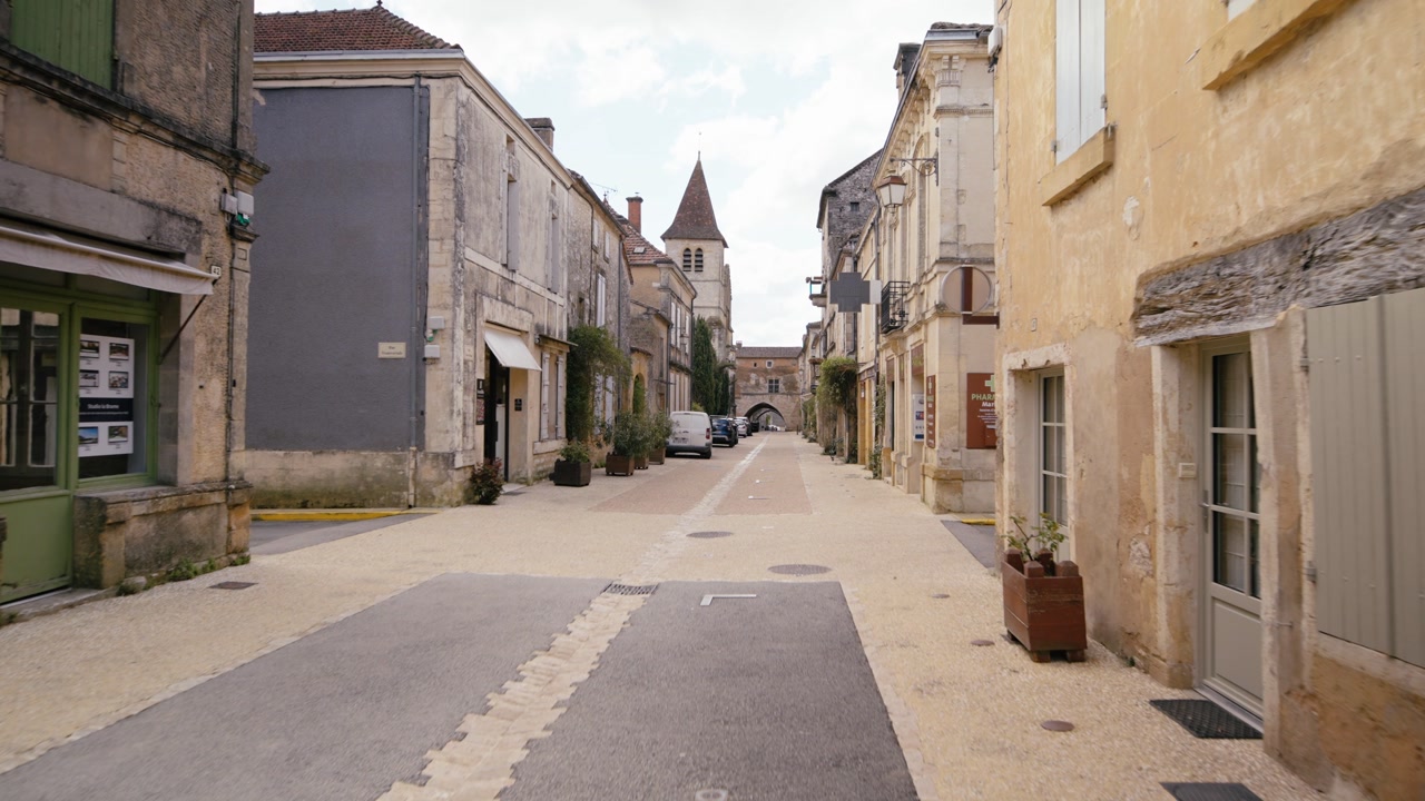 Walking through a narrow paved street in Monpazier, Dordogne, featuring traditional stone architecture and a church tower. Slow motion tracking shot.