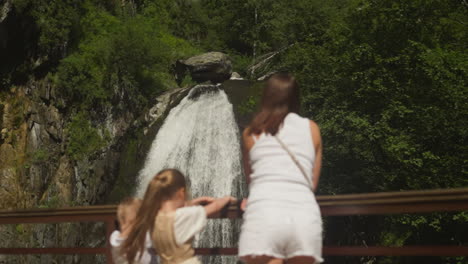 high waterfall with white sprays and happy family on bridge in wild park. woman with little children enjoy fresh cascade on mountain river in forest