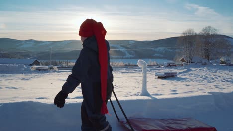 young boy wearing long red hat pulling sled across winter snow landscape in norway with bright sun on the horizon