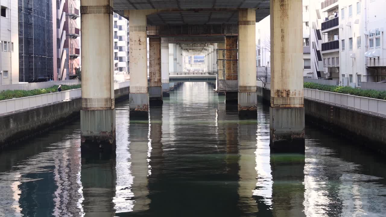 A serene view of water reflecting concrete pillars beneath a city overpass, flanked by urban buildings.