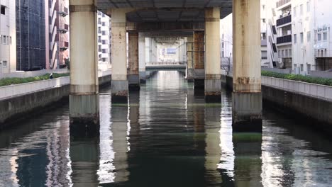 a serene view of water reflecting concrete pillars beneath a city overpass, flanked by urban buildings.