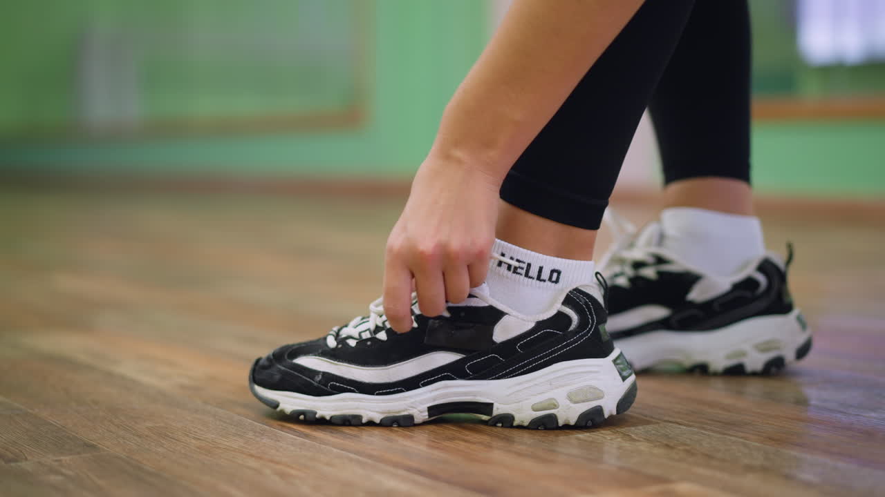 Side view of woman in black leggings tying lace of black and white sneakers while sitting on chair, wooden floor beneath creating casual active atmosphere