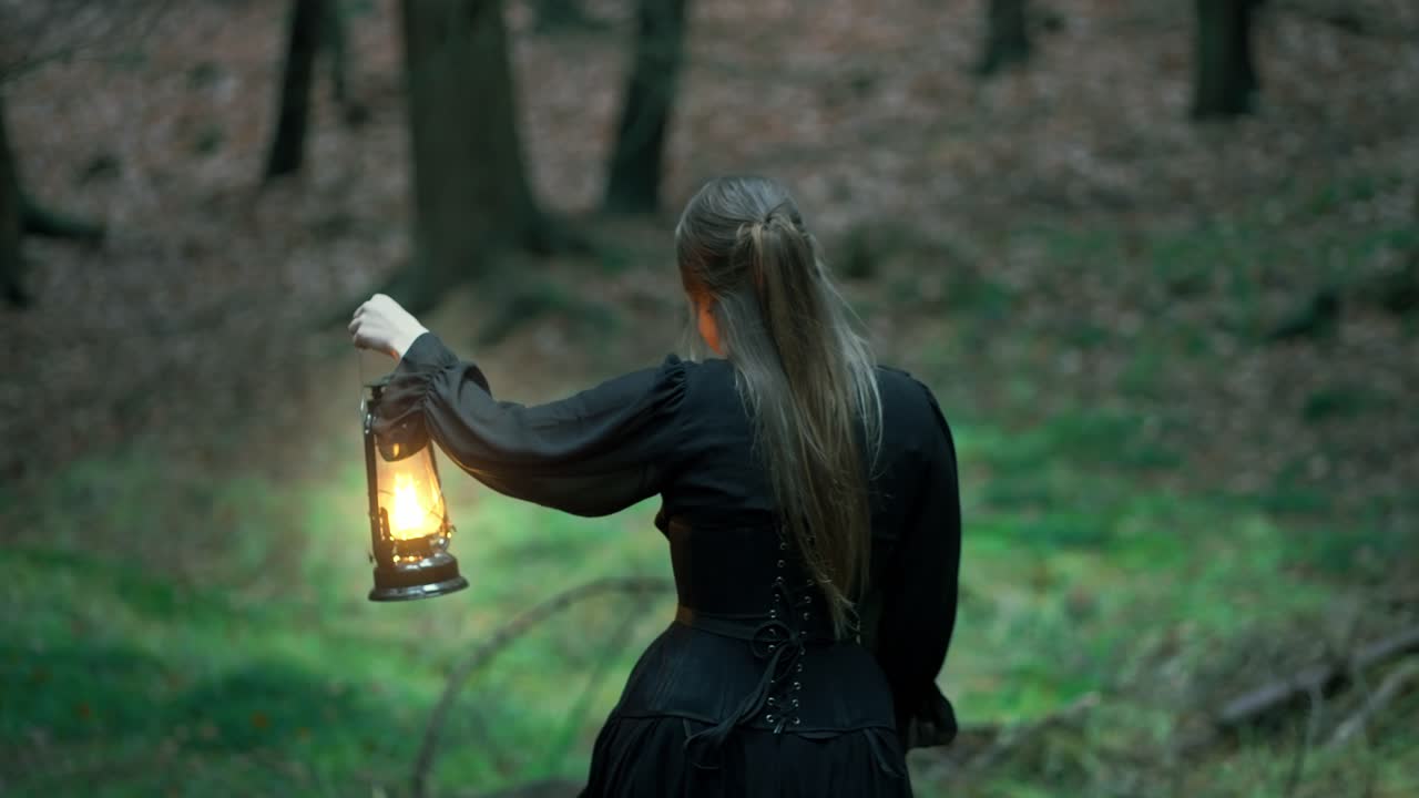 девушка с длинными волосами и черным платьем идет по темному лесу с лампой в руке