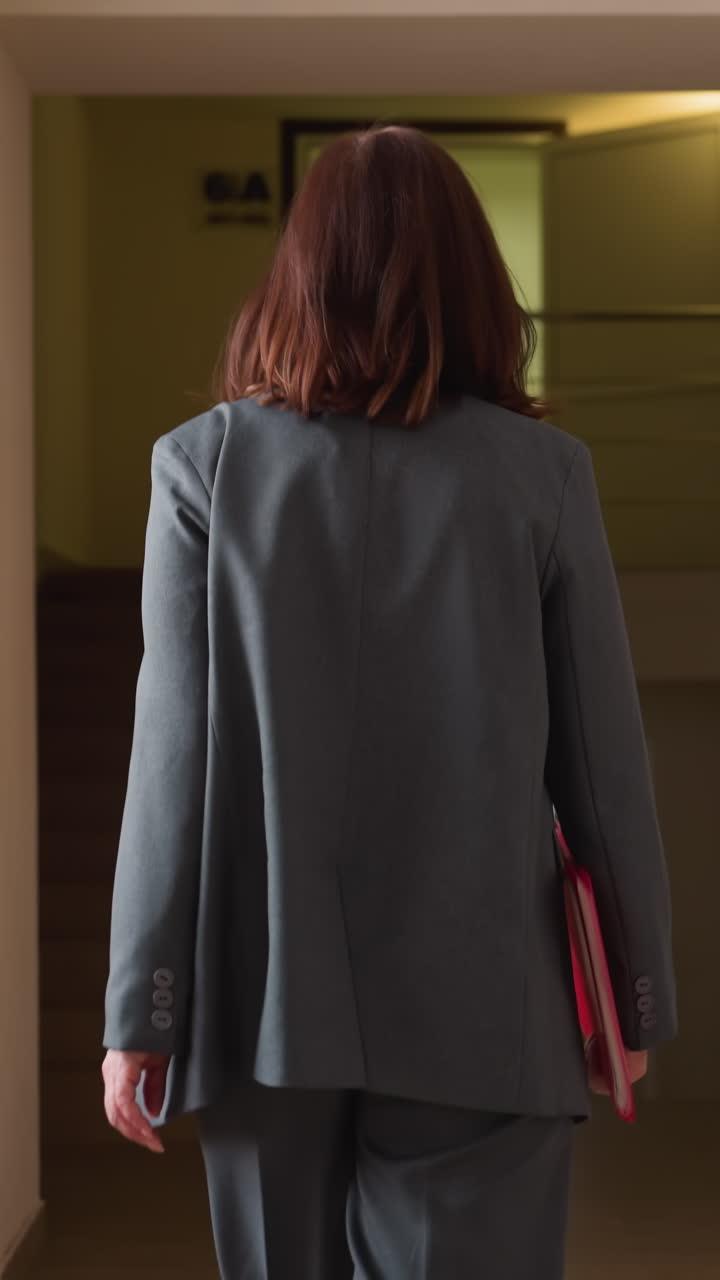 Back shot of woman walking purposefully down modern hallway, holding red folder, dressed in professional attire, exuding confidence and determination, perfect for corporate, business