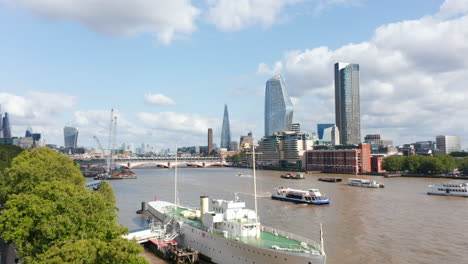 Ascending-forwards-fly-between-trees-on-waterfront.-Revealing-boats-floating-on-Thames-river.-Modern-skyscrapers-in-background.-Sunny-day-in-city.-London,-UK