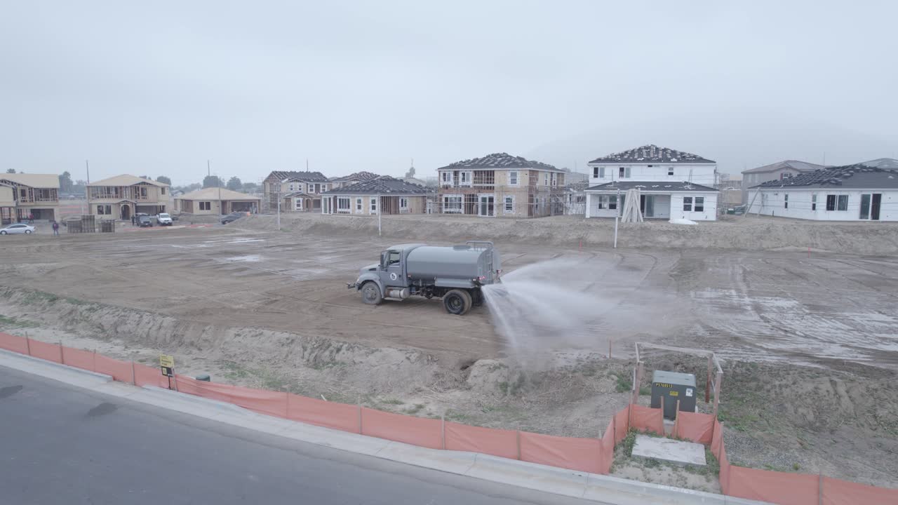 A drone observes a water truck as it diligently dumps water on a dirt lot within a home construction site, facilitating the site's preparation