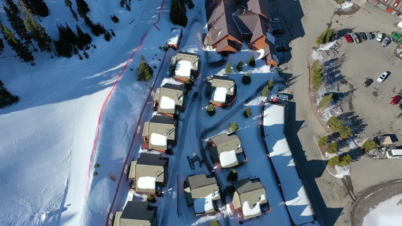 Drone glides above snowy mountain condos arranged along winding roads, with snow-draped trees and distant peaks in view, capturing a tranquil vacation rental scene in winter