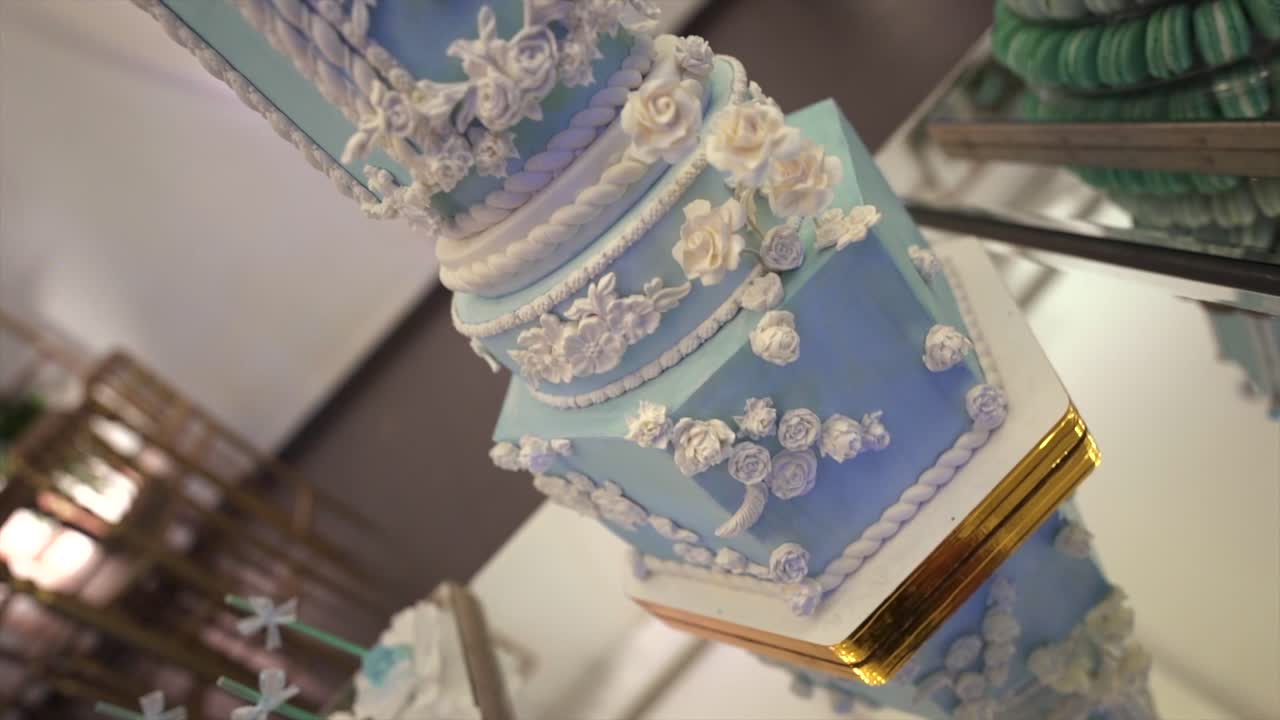 An upward camera motion reveals a tall tiered wedding cake in pastel blue with white floral and scrollwork icing, showcasing intricate craftsmanship and elegance in a formal banquet setting