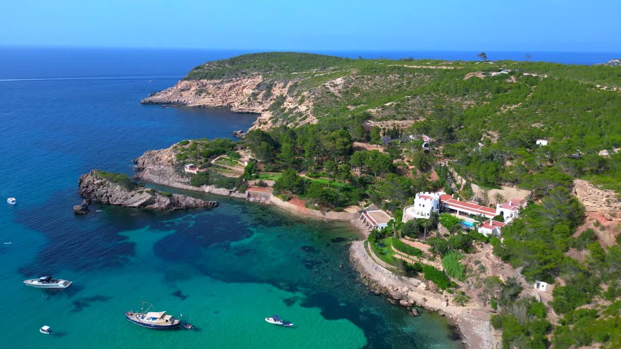 luxury villa in Ibiza, Spain, overlooking a picturesque bay with turquoise water and boats sailing on a sunny summer day. Amazing aerial view flight fly reverse drone
