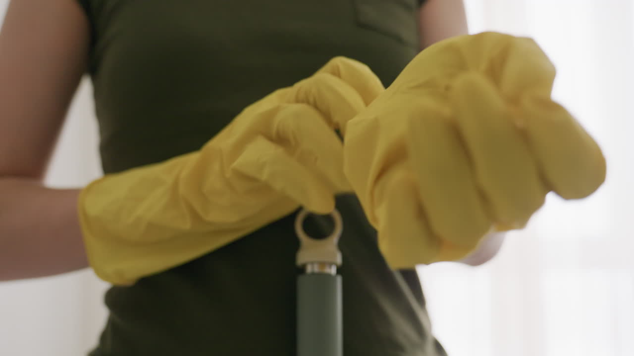 Close up of person in yellow gloves stretching fingers adjusting glove while preparing for cleaning duty indoors with mop handle resting on stomach and blurred bright background