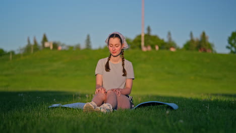 a young woman sits on her yoga mat, her glowing skin highlighted by the golden evening sun. she smiles in total well-being with a green hill behind her as the camera moves gently to the left.