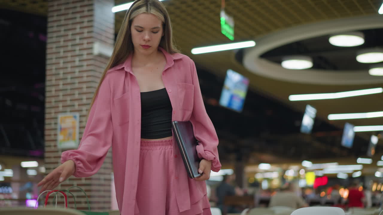 una dama blanca con un traje rosa entra en el restaurante con tres bolsas de compras en la mano derecha y una computadora portátil en la izquierda, coloca las bolsas en el asiento, se sienta y coloca la computadora portatil en la mesa