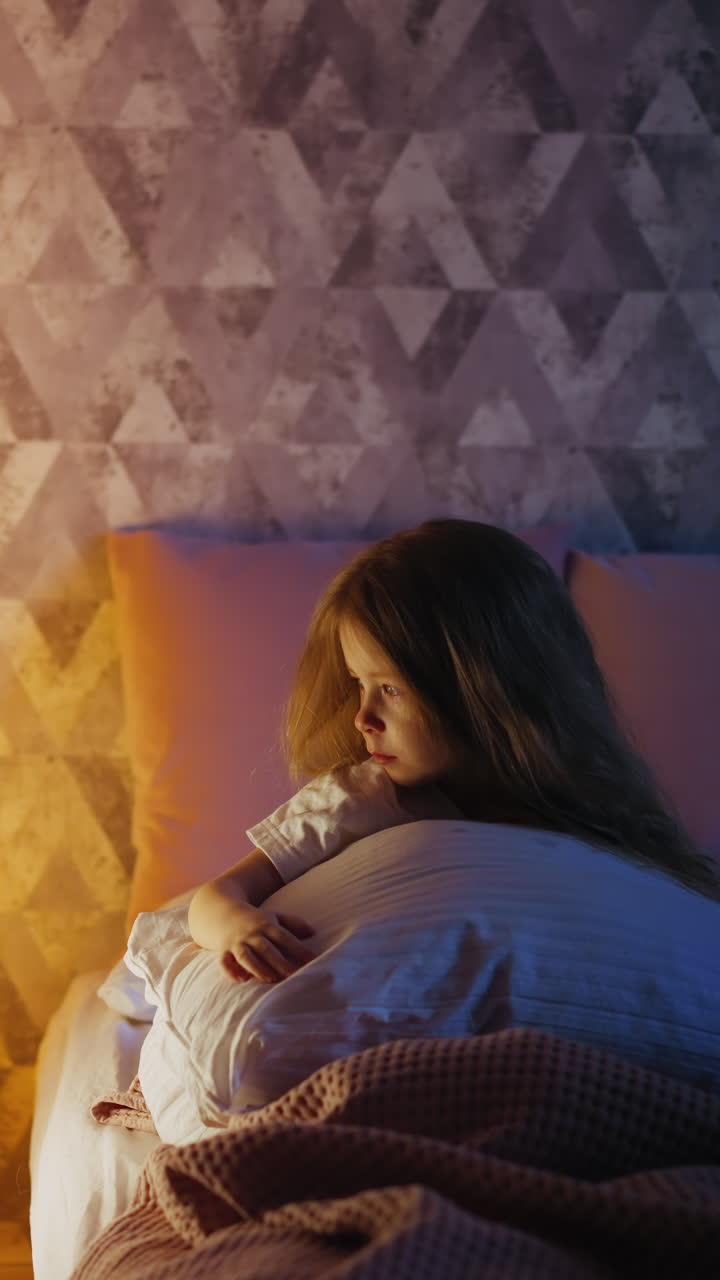 Lonely little girl with partially illuminated face hugs large pillow looking at play house with glowing night lamp on bed in cozy bedroom in evening slow motion