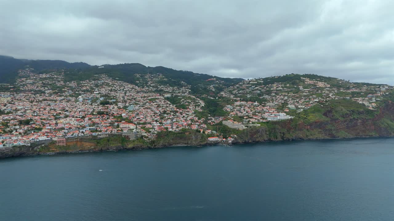 Aerial footage shows the steep hillside neighborhoods near Funchal, Madeira, with dense housing, rugged cliffs and dramatic Atlantic coastline beneath overcast skies.