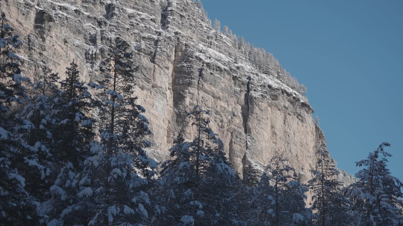 Towering cliffs and icy ridges pierce the calm sky as winter grips the forested mountain slopes. Parallax video.
