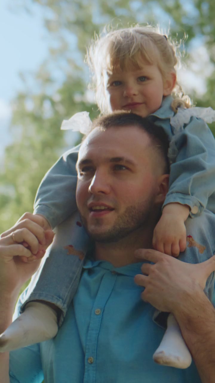 Il padre porta la sua gioiosa figlia sulle spalle. Lei ride, tirando fuori la lingua in modo giocoso. Lui sorride, godendosi il loro momento all'aperto. Il sole splende attraverso gli alberi verdi dietro di loro.