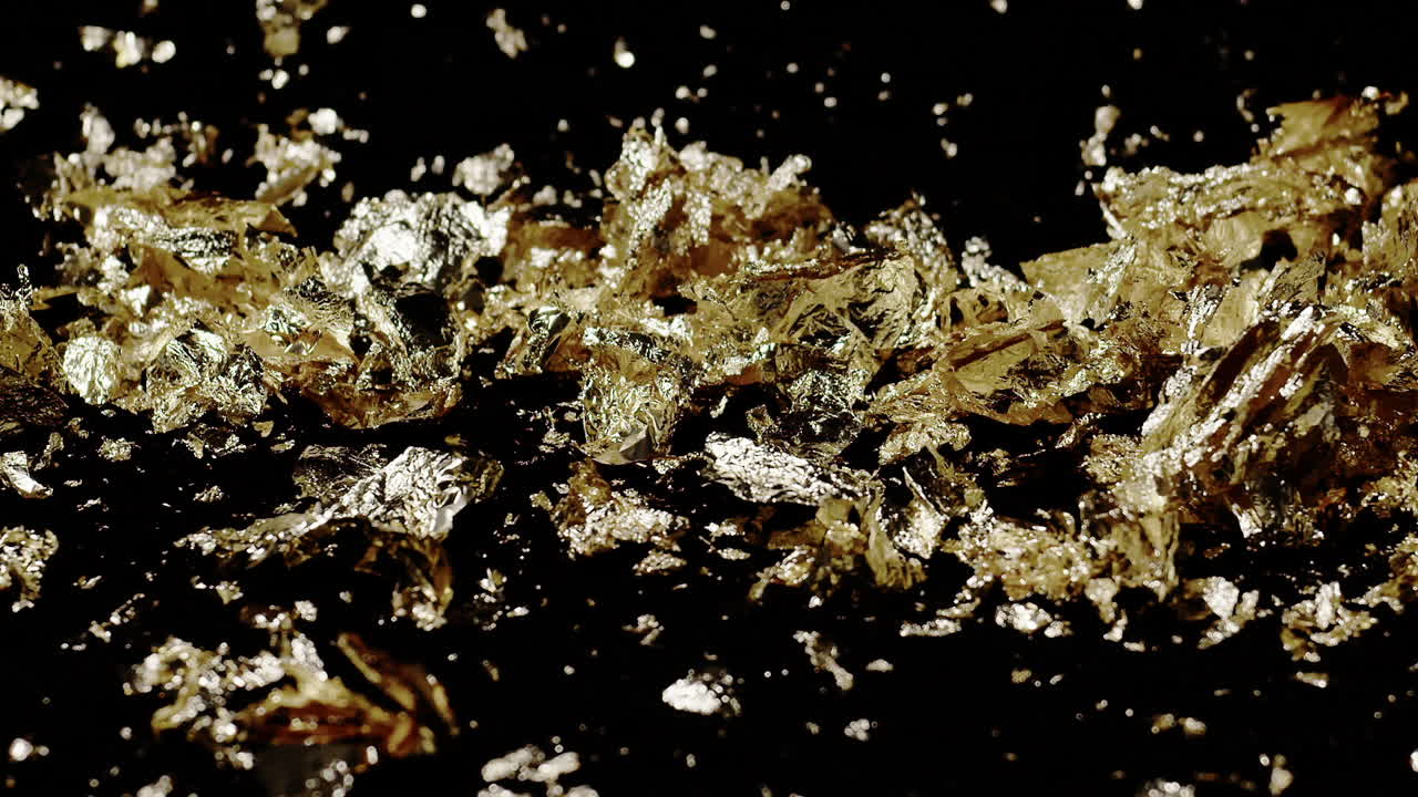 Extreme slow motion close-up of dense pile of large gold flakes blown across a black background with one blast of wind.