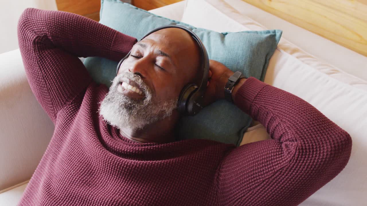 Happy senior african american man in log cabin, laying on sofa and using headphones, slow motion