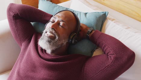 happy senior african american man in log cabin, laying on sofa and using headphones, slow motion