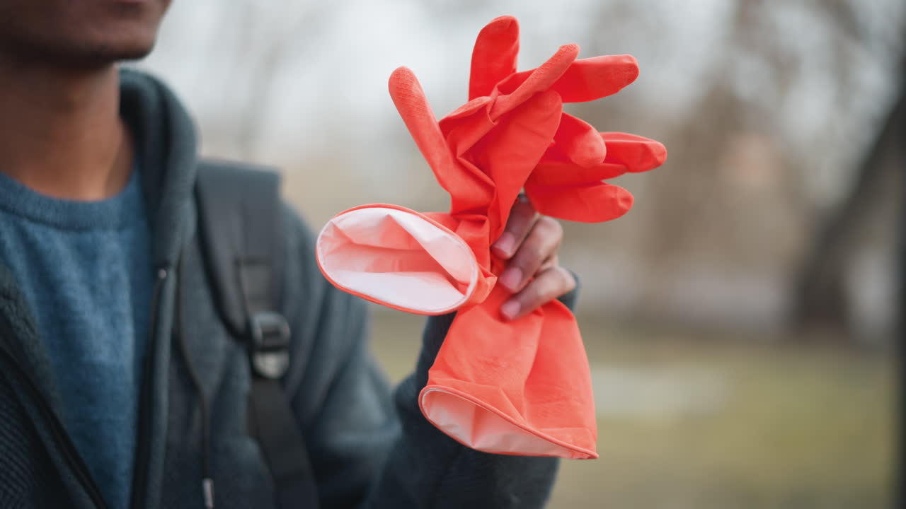 Close-up of person holding bright orange rubber gloves outdoors, wearing dark zip-up hoodie and backpack strap visible, symbolizing cleaning, volunteer work, preparation, hygiene, or outdoor community