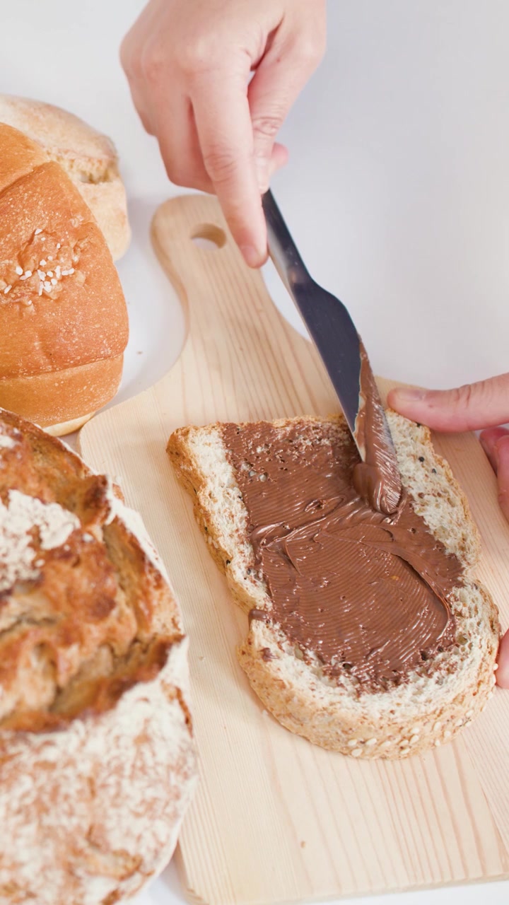 Hands use a knife to spread smooth chocolate cream on bread over a wooden board.
