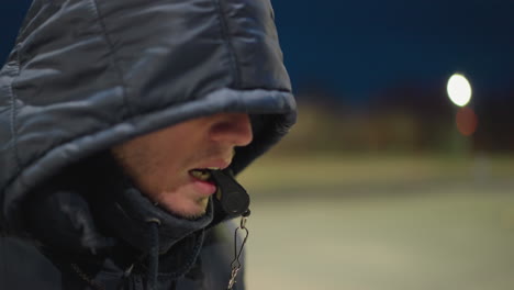 a man sits on a bench outdoors at night with a whistle in his mouth, holding a soccer ball between his legs, the background is dimly lit and blurred, featuring a distant building and a field