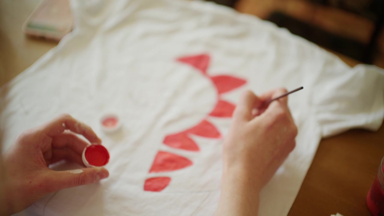 Primo piano di un artista che dipinge forme decorative rosse su una maglietta bianca usando pittura per tessuti