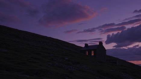 warm lights glowing from windows of a solitary house nestled on a hillside, creating a cozy scene against a backdrop of dramatic sunset hues and a cloudy sky