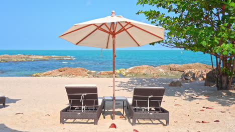 Two-empty-sun-loungers-under-a-large-canvas-shade-umbrella-on-a-white-sandy-beach-face-the-rocky-shoreline-and-incoming-tide