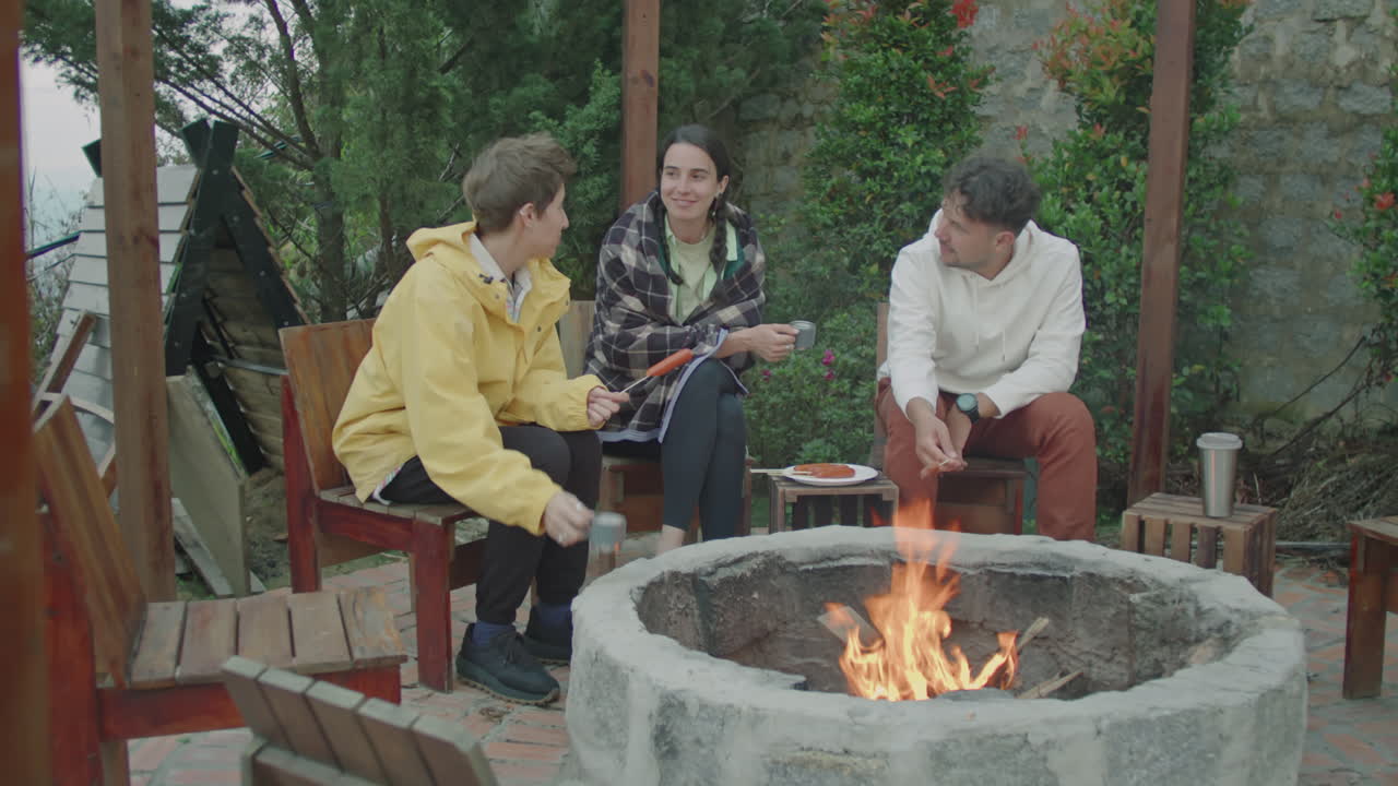 Friends Grilling Sausages and Chatting by Campfire at Glamp