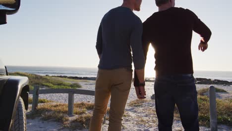 Rear-view-of-caucasian-gay-male-couple-holding-hands-and-talking-on-sunny-day-at-the-beach