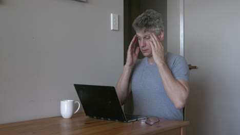 mature man wearing glasses with migraine headache working on laptop, overworked at home