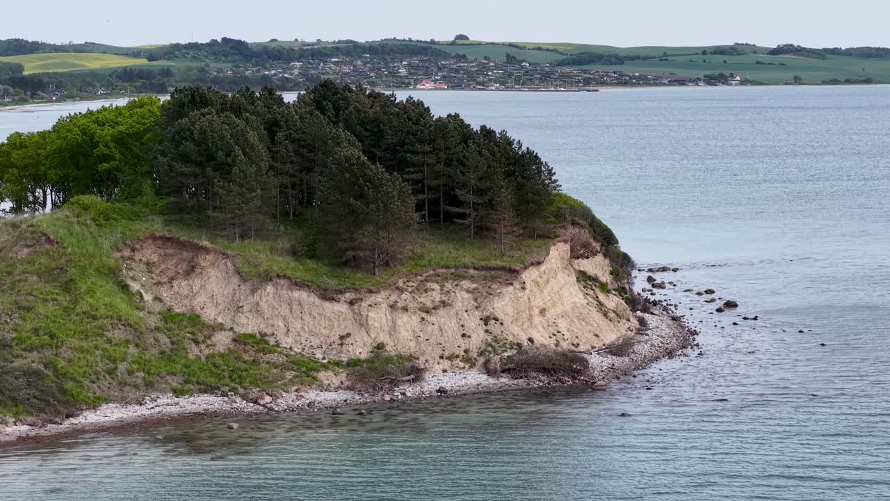 Dramatic drone footage of a coastal cliffside covered in forest, jutting into calm ocean waters with rolling hills in the background