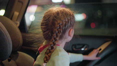 a close view of a child winding down the car glass, with a blurred view of lights across the road and passing by cars, the child has braided hair