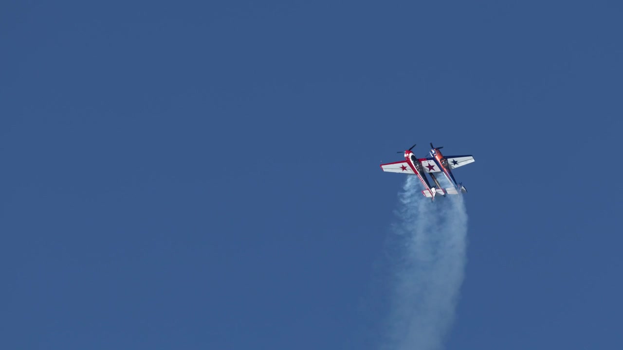 Two stunt airplanes perform synchronized aerobatic maneuvers leaving white smoke trails against clear blue sky.